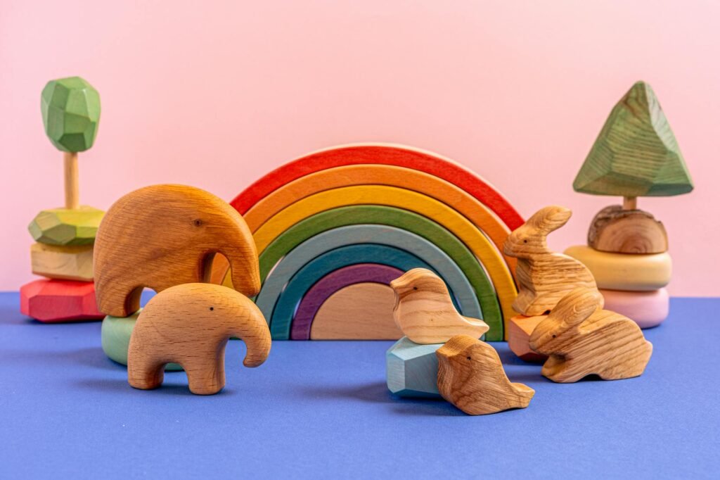 Handmade wooden Montessori toys with a colorful rainbow stacker, carved animal figures, and nature-inspired shapes on a pastel backdrop, designed to encourage toddler sensory play and open-ended learning.